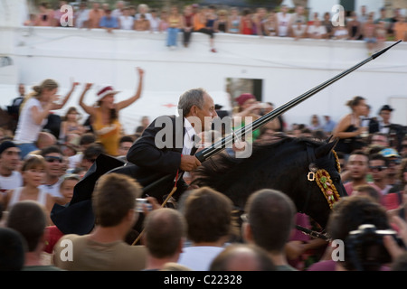 Un cavallo e cavaliere tenendo una lancia carica attraverso una folla di persone nella città di Ciutadella durante la Festa de Sant Joan in Menorca. Foto Stock