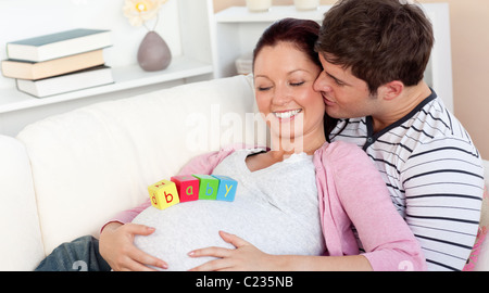 Giovane uomo baciare sua moglie incinta con il bambino lettere sul ventre sdraiato sul divano Foto Stock