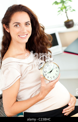 Gravidanza radiante imprenditrice tenendo una sveglia e seduto alla sua scrivania Foto Stock