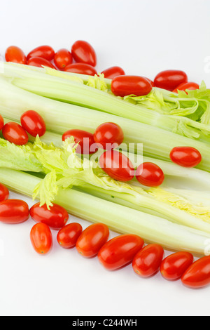 Un sacco di piccoli pomodori rossi con organici cuore di sedano gambi sotto. Lungo fresco vegetali verdi e piccoli rosso ovale mix di frutta su bianco in corrispondenza di un angolo. Foto Stock