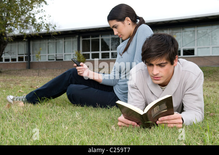 Gli studenti universitari che giace sul campo guardando un telefono cellulare e la lettura di un libro Foto Stock