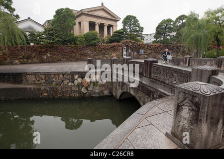 Il ponte di pietra che attraversano il Canal Bikan nel quartiere storico con museo d'Arte Ohara in background, Kurashiki, Giappone. Foto Stock