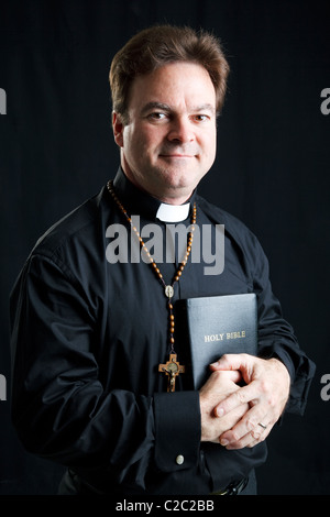 Ritratto di un sacerdote con un rosario e una Bibbia. Illuminazione drammatica su sfondo nero. Foto Stock