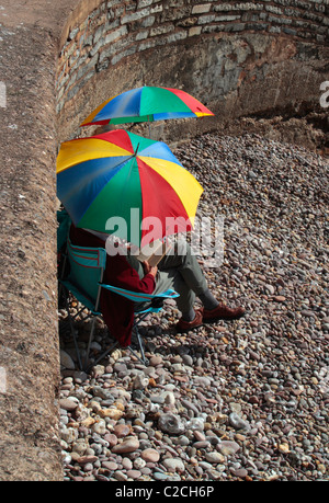 Coppia inglese a sedersi su una spiaggia in Devon utilizzando ombrelloni colorati Foto Stock