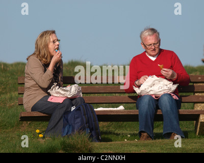 Vecchia coppia seduto su una panchina a mangiare pesce e patatine, Cornwall, Regno Unito Foto Stock