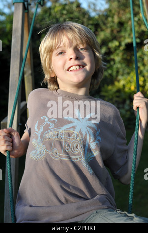 Biondo ragazzo giocando su un giardino swing sorridente e tenere le funi in estate il sole Foto Stock
