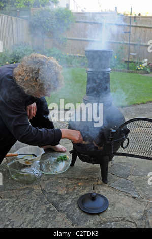 Donna che utilizza una ghisa Giardino Chimenea per cucinare e barbeque cibo barbecue nel giardino estivo del Regno Unito Foto Stock