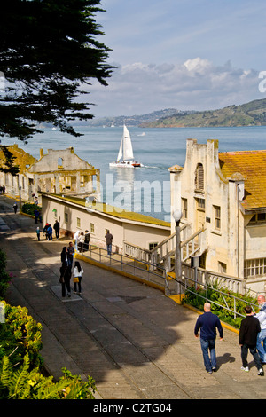 I turisti a piedi passato un deterioramento degli edifici di stoccaggio presso l'ex Alcatraz prigione federale nella Baia di San Francisco, CA. Nota barca a vela Foto Stock