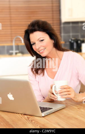 Donna che utilizza il suo computer portatile in cucina tenendo una tazza di tè o caffè Foto Stock