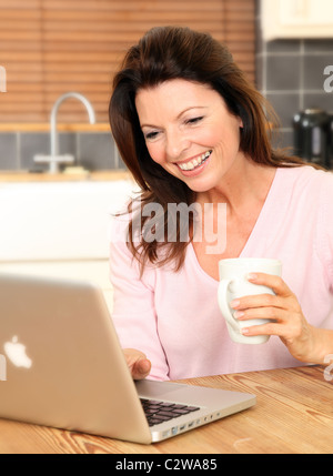 Donna felice con il suo computer portatile in cucina tenendo una tazza di tè o caffè Foto Stock