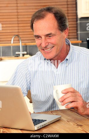 Uomo felice usando il suo computer portatile in cucina tenendo una tazza di tè o caffè Foto Stock