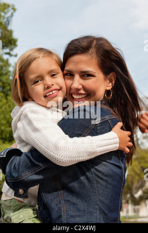 Bella sorridente madre ispanica in jean Jacket avvolge il suo felice giovane figlia che indossa un maglione bianco e restituisce il suo abbraccio Foto Stock