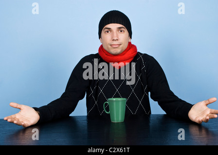 Giovane con vuota espressione seduta a tavola con una tazza di una bevanda calda,vestito in abiti invernali e mi chiedo che cosa sta succedendo Foto Stock