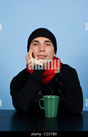 Giovane uomo triste in abbigliamento invernale in piedi a tavola con la mano al mento e riflettendo su sfondo blu Foto Stock