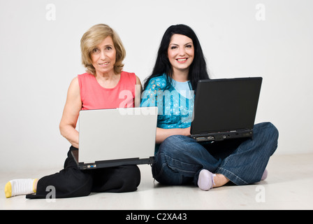 Madre e figlia seduti con le gambe incrociate sul pavimento in legno in casa e sia tramite computer portatile e sorridente Foto Stock