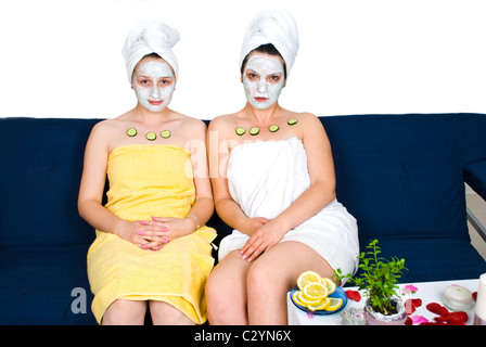 Due giovani amici di donna con maschere facciali e fette di cetriolo sulla loro pelle seduta sul lettino in una spa salone Foto Stock