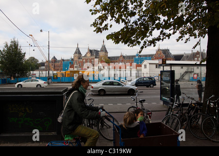 Una donna peddles lontano con due bambini animati ingannare intorno al carrello anteriore di fronte la Stazione Centrale di Amsterdam Foto Stock