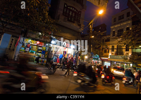 In orizzontale ampia angolazione di negozi e ciclomotori, un tipico streetscene del vecchio quartiere illuminata di notte, nel centro di Hanoi. Foto Stock