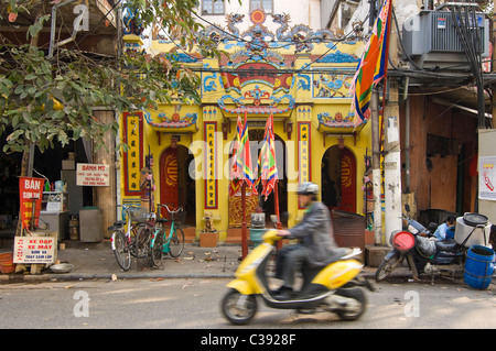 Vista orizzontale di un piccolo tempio schiacciata tra le case nel vecchio quartiere nel centro di Hanoi con una guida di scooter passati. Foto Stock