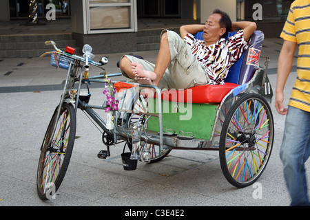 Uomo dorme sul suo coloratissimo taxi bicicletta su una strada di Singapore Foto Stock