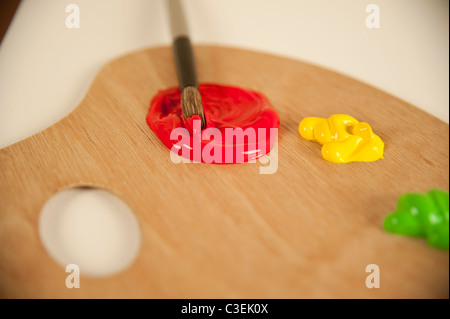 Un pennello è immerso nel colore rosso brillante pittura ad olio su una tavolozza di legno, contro una tela vuota in un studio d'arte. Foto Stock