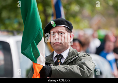 Uomo in un stile paramilitari mantiene uniforme un tricolore irlandese come parte di un partito di colore alla testa di una parata repubblicana. Foto Stock