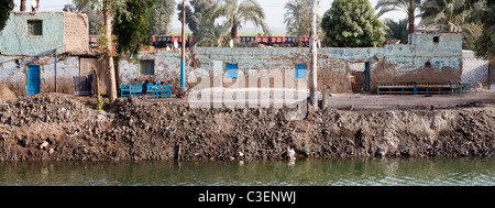 Tradizionale egiziana case lungo un canale fangoso banca, con banchi di blu e i bambini nella parte anteriore e di un treno di merci dietro, Egitto Foto Stock
