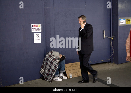 Imprenditore sul suo telefono cellulare passa da una verso il basso e fuori dalla giovane uomo a mendicare per strada per aiuto monetario. La città di New York. Foto Stock