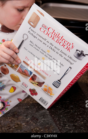 Giovane ragazza in cucina che indossa Tracy Beaker grembiule, tenendo, leggendo & concentrandosi sulla ricetta nel libro di cucina dei bambini di Dorling Kindersley - Inghilterra, Regno Unito. Foto Stock