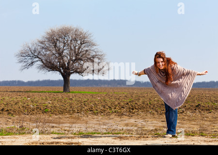 Felice redhead giovane donna in un campo con un grande albero in background Foto Stock