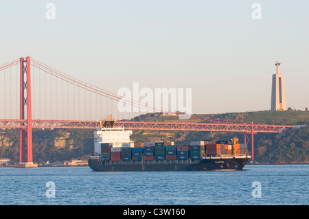 Una nave a pieno carico con contenitori passando sotto il Ponte 25 de Abril sul fiume Tago a Lisbona, Portogallo. Foto Stock