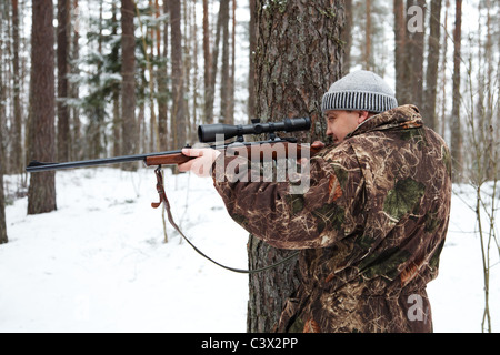 Hunter di camuffamento di mira con il fucile da cecchino a foresta d'inverno. Foto Stock