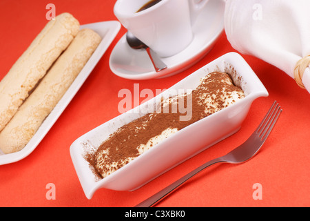 Il Tiramisù' torta servito su una piastra bianca. Savoiardi e tazza di caffè al di fuori della messa a fuoco in background Foto Stock