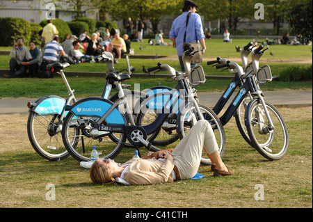 Una donna sunbaths in un parco di Londra vicino a Barclays Cycle biciclette a noleggio Foto Stock