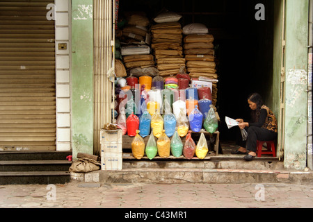 Colorato / colorate sacchetti di polvere di vernice o pigmenti in vendita su strada di vernice ad Hanoi, Vietnam. Donna venditore legge giornale Foto Stock