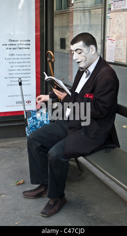 Uomo vestito in costume da clown seduta a una fermata di autobus e leggere un libro mentre è in attesa per un autobus Foto Stock