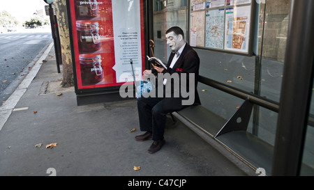 Uomo vestito in costume da clown seduta a una fermata di autobus e leggere un libro mentre è in attesa per un autobus Foto Stock