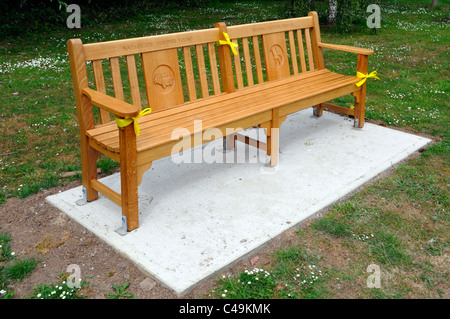 Nuovo legno ampio parco pubblico sedile unico sul pavimento di cemento nastro giallo iscrizione e pannelli scolpiti posti come memoriale sulla verde villaggio Essex England Regno Unito Foto Stock