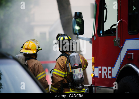 Due vigili del fuoco in piedi accanto a firetruck osservando il fumo offuscato il fuoco. Città Branding specifico rimosso dal motore Fire. Foto Stock
