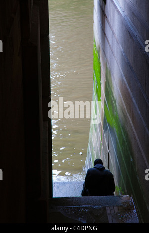 Un uomo seduto sui gradini che conducono in basso verso il fiume Tamigi sotto il ponte di Londra, London, Regno Unito Foto Stock