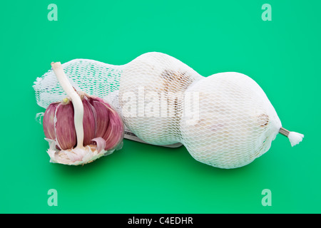 Due interi bulbi di aglio e una metà che mostra i chiodi di garofano in una borsa a rete su un verde tagliere Foto Stock