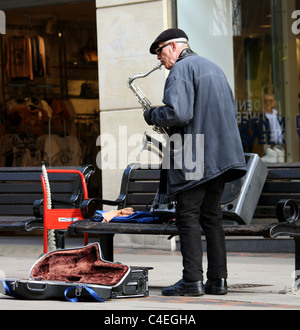 Un uomo anziano musicista di strada sul lato della strada a fianco di una panca a giocare il suo sassofono al di fuori di un negozio di abbigliamento, musica blues per denaro Foto Stock