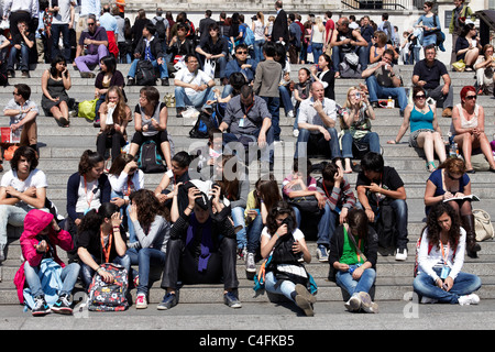 Turisti e londinesi seduti sui gradini in Trafalgar Square e godetevi il sole sulla il giorno più caldo dell'anno finora Foto Stock