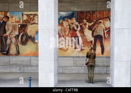 Una donna che guarda la parete socialista murale di felice i lavoratori sulla parete del ministero delle Finanze in edificio ex Berlino Est Foto Stock