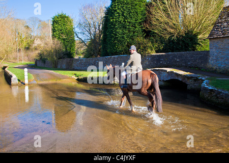 Un cavallo e cavaliere attraversando la Ford nel grazioso villaggio Costwold di Shilton, Oxfordshire, Inghilterra, Regno Unito in una giornata di sole in primavera Foto Stock