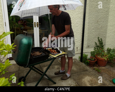 Uomo con ombrello e salsicce di cottura su un Barbeque sotto la pioggia nel giardino Inghilterra Foto Stock