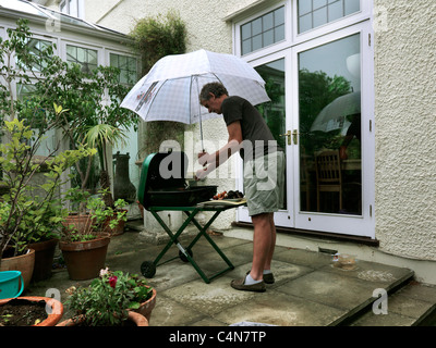 Uomo con ombrello e salsicce di cottura su un Barbeque sotto la pioggia nel giardino Inghilterra Foto Stock