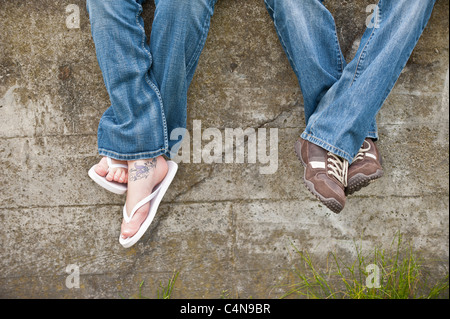 Due coppie di gambe in jeans da una giovane coppia penzolare contro un muro di cemento al di fuori Foto Stock