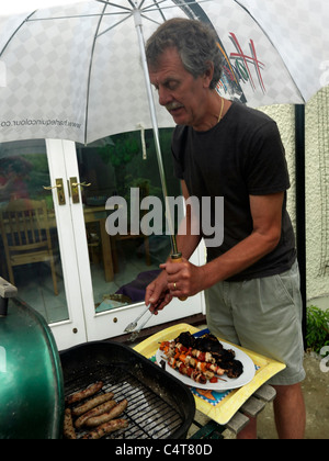 Uomo con ombrello e salsicce di cottura su un Barbeque sotto la pioggia nel giardino Inghilterra Foto Stock