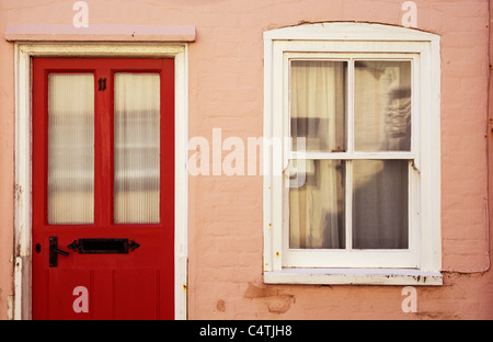 Dettaglio della casa rosa con pareti dipinte di rosso porta anteriore e sash bianco finestra con tendina inondata dalla calda luce del sole riflessa Foto Stock